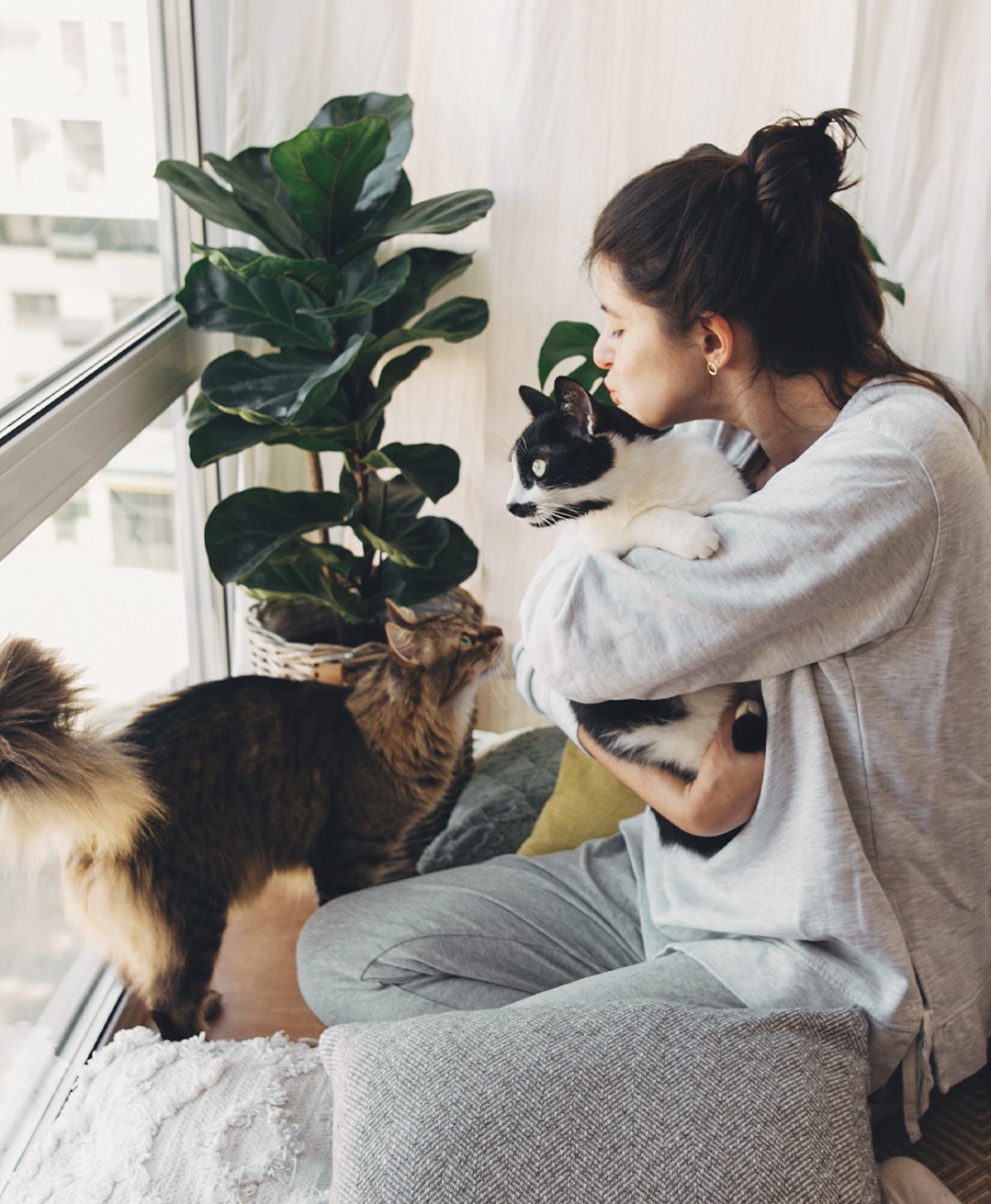 A young woman holding and kissing one of her cats while another cat looks up lovingly at her